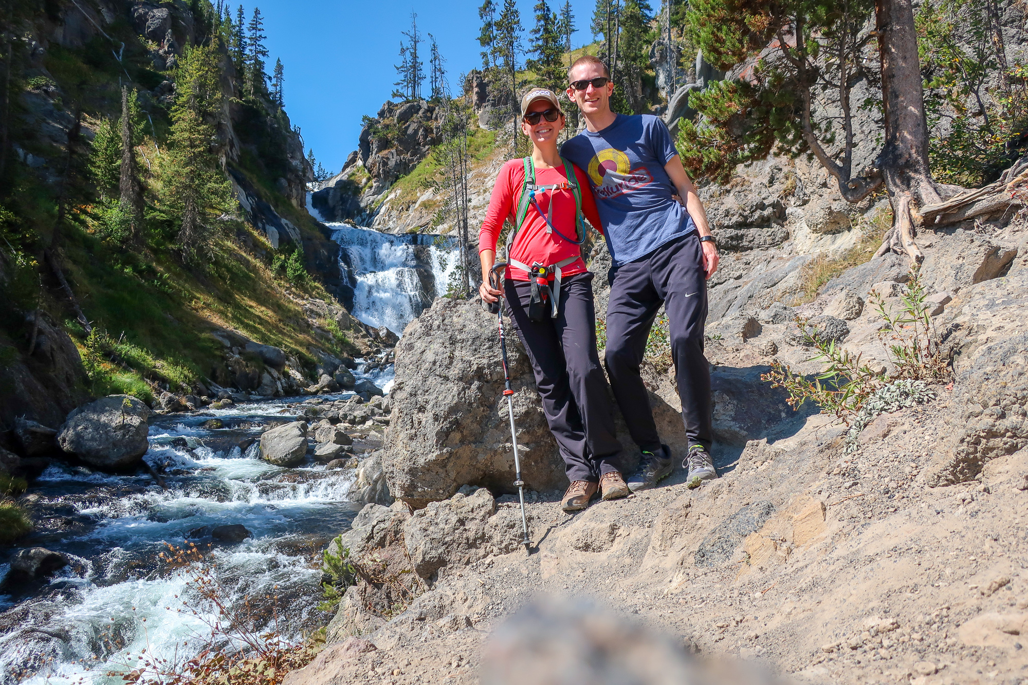a couple in hiking clothes pose for a picture in front of mystic falls, a large waterfall in yellowstone national park